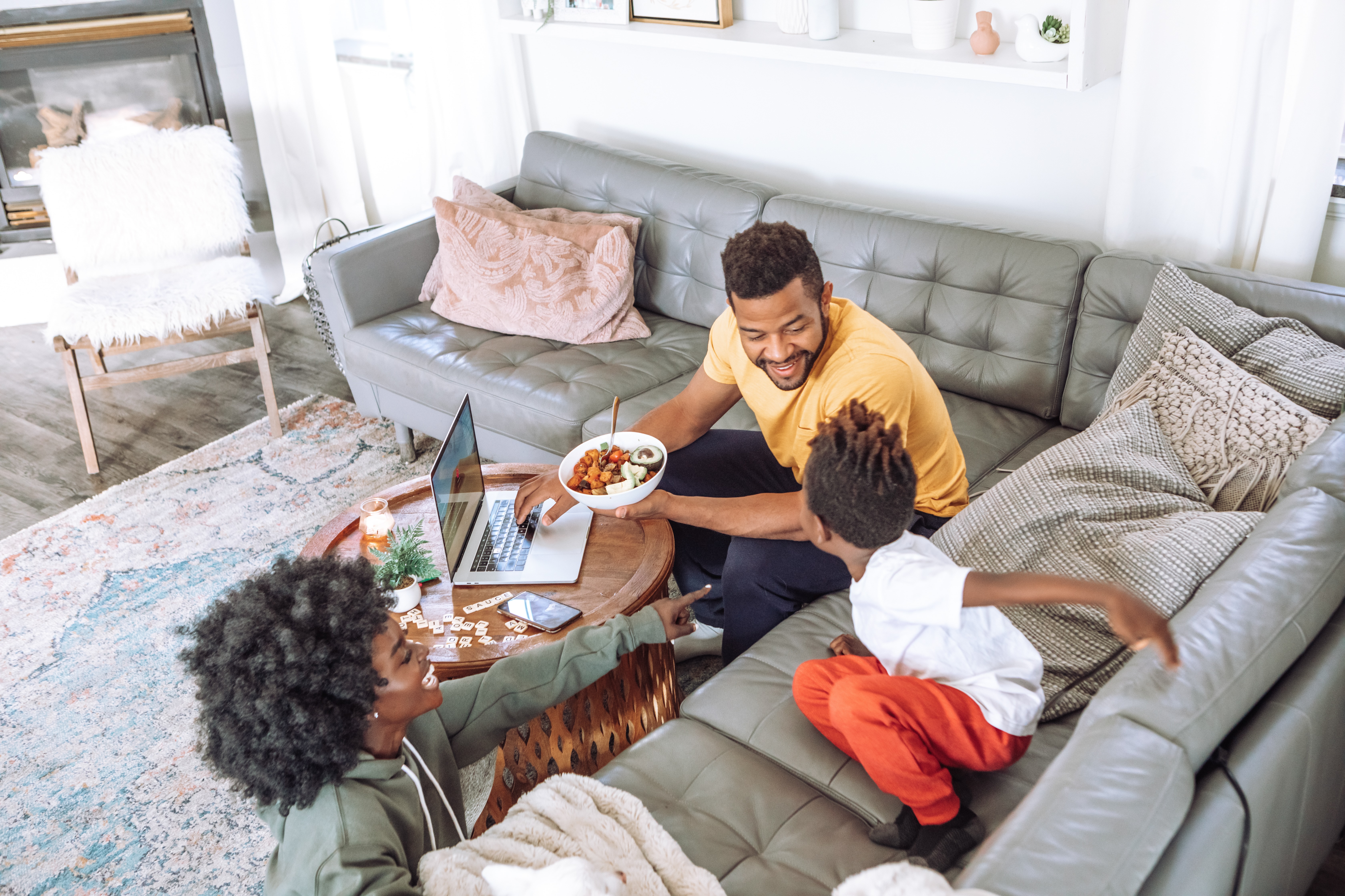 Three Black people sitting in a living room. One of the people is a child crouched on a couch. The two older people are engaging with the child.
